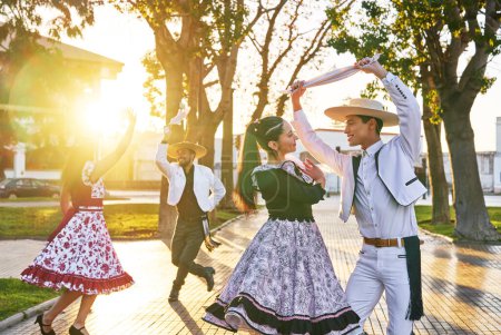 Foto de Grupo de cuatro jóvenes latinoamericanos vestidos de huaso bailando cueca en la plaza del pueblo al atardecer - Imagen libre de derechos