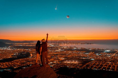 Foto de Siluetas de una pareja de pie en la cima de la colina mirando la noche estrellada con Luna y estrella fugaz en el cielo sobre la luz de la ciudad - Imagen libre de derechos