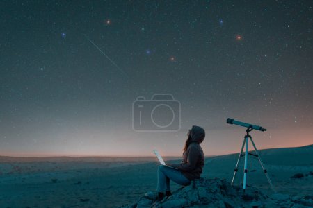  woman sitting in the desert with a laptop next to a telescope at night watching the starry sky, astronomy and stargazing concep