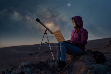 Frau sitzt in der Wüste mit einem Laptop neben einem Teleskop in der Nacht unter den Sternen und Milchstraße, Astronomie und Sternenguckkonzept 