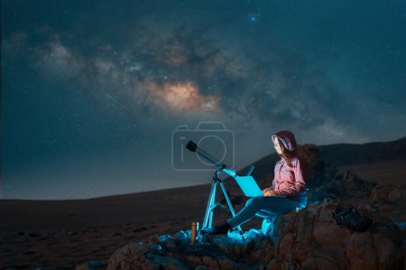 Digitale Nomadenfrau, die nachts in der Wüste mit Laptop neben einem Teleskop unter Sternen und Milchstraße sitzt, Astronomie und Sternenbeobachtungskonzept