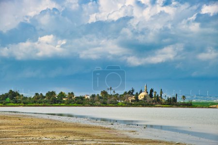 Hala Sultan Tekke, or the Mosque of Umm Haram, on the bank of Larnaca Salt Lake, Cyprus, under dark thunderclouds. Wind turbines on the hills in the background.