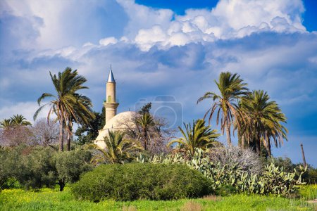The Ottoman Turkish Hala Sultan Tekke, or Mosque of Umm Haram (1760), in Larnaca, Cyprus, surrounded by tall palm trees and cacti, under dark thunderclouds. 