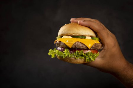 Hand holding a cheeseburger on dark background.