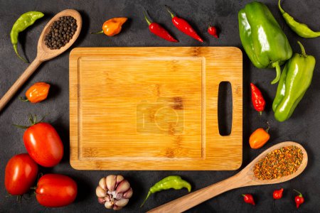 Foto de Tabla de cortar vacía en la mesa con ingredientes para cocinar. Tomates, varios pimientos, ajo y pimientos verdes. - Imagen libre de derechos