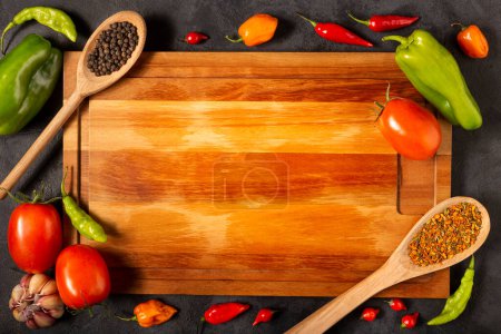 Foto de Tabla de cortar vacía en la mesa con ingredientes para cocinar. Tomates, varios pimientos, ajo y pimientos verdes. - Imagen libre de derechos