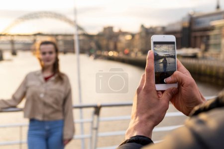 Photo for A young couple spending the day in Newcastle Upon Tyne. The woman is posing for a photo her boyfriend is taking on a mobile phone while she stands on a bridge with the River Tyne behind her. - Royalty Free Image