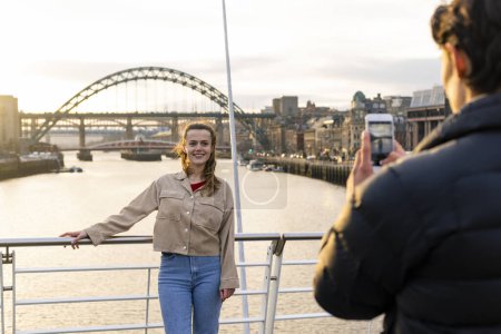 Photo for A young couple spending the day in Newcastle Upon Tyne together. The woman is posing for a photo her boyfriend is taking on a mobile phone while she stands on a bridge with the River Tyne behind her. - Royalty Free Image