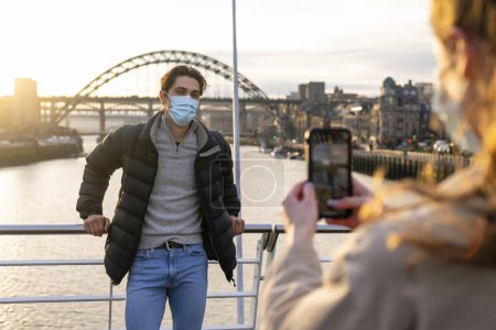 Photo for A young couple spending the day in Newcastle. One man is posing for a photo his girlfriend is taking on a mobile phone with the River Tyne behind him. They are wearing protective face masks. - Royalty Free Image