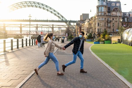 Photo for A young couple spending the day in Newcastle Upon Tyne. They are holding hands and walking near the River Tyne while the man looks back at his girlfriend. They are wearing face masks. - Royalty Free Image