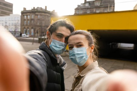 Photo for A young couple spending the day in Newcastle Upon Tyne. They are standing on a paved area in the city centre while taking a selfie. They are looking at the camera and wearing protective face masks. - Royalty Free Image