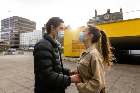 Foto de Una joven pareja pasando el día en Newcastle Upon Tyne. Están parados en una zona pavimentada en el centro de la ciudad mientras se miran y se toman de la mano. Llevan máscaras protectoras. - Imagen libre de derechos