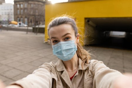 Photo for A young woman spending the day in Newcastle Upon Tyne. She is standing on a paved area in the city centre while taking a selfie. She is looking at the camera and wearing a protective face mask. - Royalty Free Image