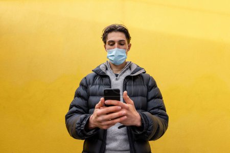 Photo for A young man spending the day in Newcastle Upon Tyne. He is standing against a yellow wall and using his phone to text a friend. He is wearing protective face mask. - Royalty Free Image