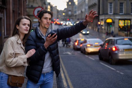 Foto de Una joven pareja pasando el día en Newcastle Upon Tyne juntos. Están de pie junto a una carretera y el hombre está tratando de llamar a un taxi mientras ambos miran a la distancia. - Imagen libre de derechos