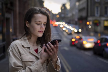 Photo for A young woman standing on a city street in Newcastle Upon Tyne in the evening while talking to a friend on her mobile phone. She is waiting on a taxi. - Royalty Free Image