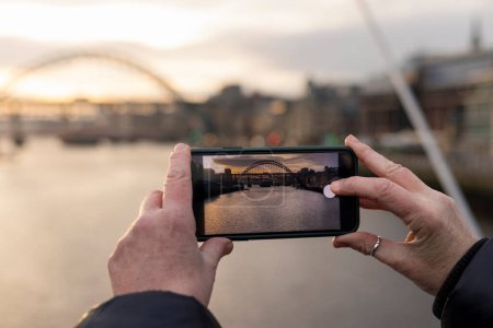 Photo for An unrecognisable woman taking a photo with a mobile phone of the River Tyne at dusk in Newcastle Upon Tyne. The bridges can be seen in the photograph. - Royalty Free Image