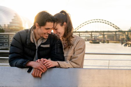 Photo for A young couple spending the day in Newcastle Upon Tyne together. They are standing on a bridge with the river tyne and other bridges behind them while embracing each other and looking happy. - Royalty Free Image