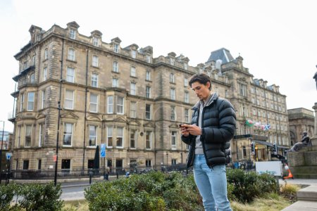 Photo for A wide-view shot of a young man standing in the city centre, he is using his smartphone to send a text message. - Royalty Free Image