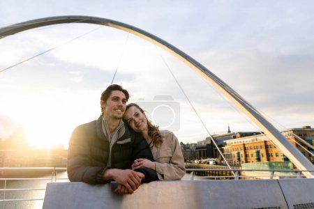 Photo for A wide-view shot of a young couple standing side by side looking at the view together on a bridge in the city, they are in love. - Royalty Free Image
