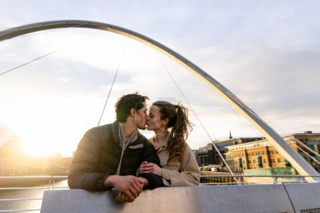Photo for A wide-view shot of a young couple standing side by side sharing a kiss together on a bridge in the city, they are in love. - Royalty Free Image