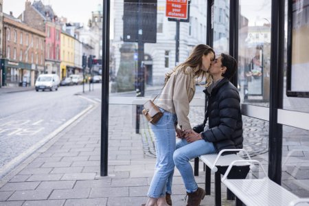 Photo for A side-view shot of a young couple waiting at a bus stop together, they are in love and sharing a kiss. - Royalty Free Image