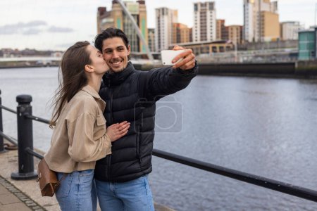 Photo for A wide-view shot of a young couple standing together kissing and taking a selfie, they are in love. - Royalty Free Image