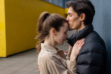 Photo for A side-view shot of a young couple standing face to face together against a wall, they are embracing and sharing a kiss, they're in love. - Royalty Free Image