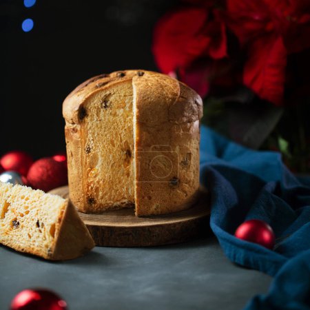 Foto de Un pedazo de pan dulce panettone, comida italiana tradicional para Navidad sobre un fondo oscuro con luces, acebo y guirnalda en la mesa, de cerca - Imagen libre de derechos