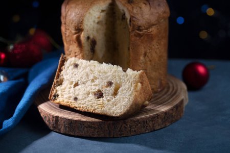 Téléchargez les photos : Un morceau de pain sucré panettone avec des raisins secs, cuisine italienne traditionnelle pour Noël sur un fond sombre avec des lumières, houx et guirlande sur la table - en image libre de droit