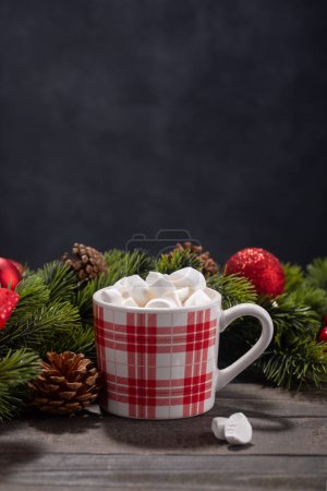 Téléchargez les photos : Red mug with hot chocolate on top of which pieces of marshmallows float, and behind are branches of a Christmas tree decorated with red balls, with copy space on wooden background - en image libre de droit