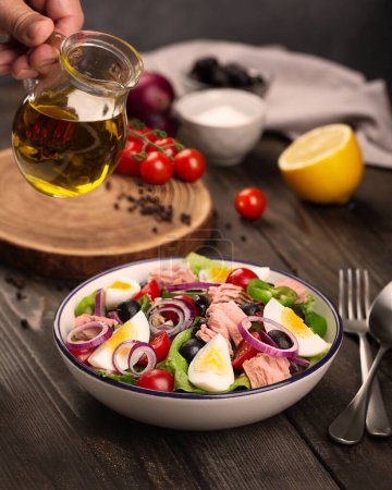 Téléchargez les photos : Nicoise salad with tuna, egg, olives and lettuce leaves on a wooden table. A mans hand holds a small jug of olive oil. High quality photo - en image libre de droit