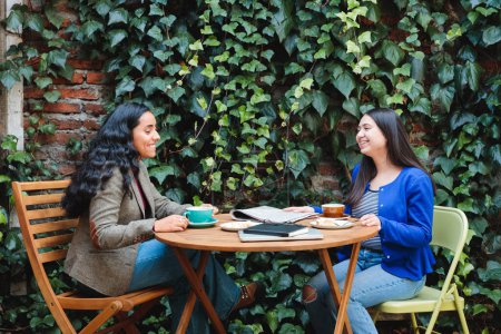 Téléchargez les photos : Amis et café : Vibrant Café Patio accueille deux jeunes femmes latines appréciant le café et délicieuses pâtisseries sucrées. Photo de haute qualité - en image libre de droit