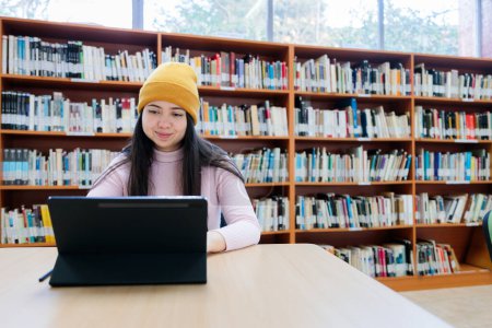 Téléchargez les photos : Poursuites académiques : Jeune étudiante universitaire étudiant avec sa tablette à la bibliothèque. Photo de haute qualité - en image libre de droit