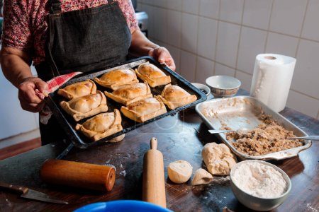 Téléchargez les photos : Cuisine traditionnelle chilienne : femme latina méconnaissable tenant un plateau d'empanadas cuits au four dans sa cuisine rustique. Photo de haute qualité - en image libre de droit