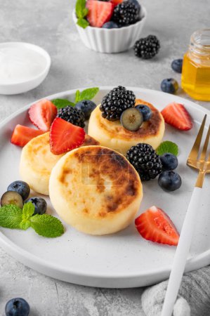 Cottage cheese pancakes, syrniki, ricotta fritters with fresh berries, honey and sour cream on a gray concrete background. Healthy and delicious breakfast. Top view, copy space