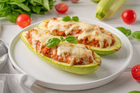 Zucchini boats stuffed with meat, vegetables and cheese on a white plate on a white wooden background