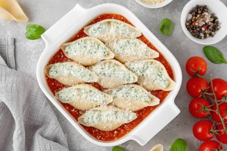 Raw conchiglioni pasta stuffed ricotta cheese and spinach with tomato sauce and parmesan cheese on top in a white baking dish on a gray concrete background