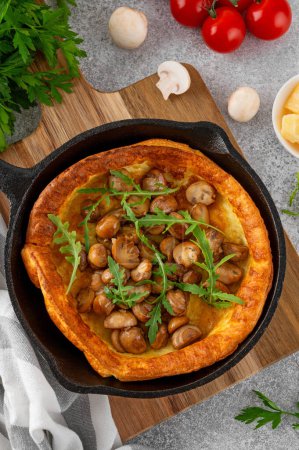 Savory Dutch baby pancake with cheese, fried mushrooms and arugula in cast iron pan on a concrete background. Vegetarian breakfast