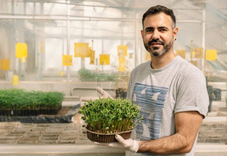Foto de Male researcher smiles and looks at camera, holding soybean sprout plant in greenhouse. - Imagen libre de derechos