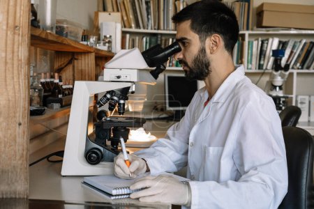 Foto de Male scientist looking under microscope, analyzes sample in petri dish. Biotechnological research in laboratory. - Imagen libre de derechos