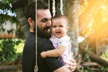 Foto de Retrato del padre y el bebé sonriendo y mirando a la cámara sentada en una hamaca. Concepto del Día del Padre. Luz del atardecer - Imagen libre de derechos