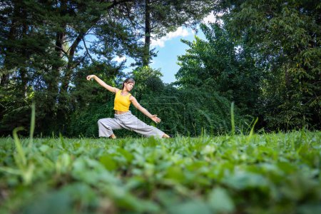 Foto de Mujer haciendo ejercicio qi gong tai chi. Una mujer practica tai chi en la naturaleza rodeada de árboles y un cielo azul. Primer plano de la hierba. - Imagen libre de derechos