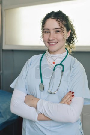 Foto de Enfermera médica joven sonriente vestida con bata blanca y estetoscopio alrededor del cuello parada en una clínica privada con los brazos cruzados mirando a la cámara. Retrato vertical. - Imagen libre de derechos