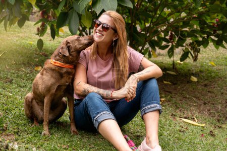 La mujer feliz con gafas de sol y su perro sentado en el parque, su mascota le da un beso en la mejilla.