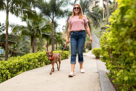 Foto de Una mujer de mediana edad sonríe y mira a la cámara mientras camina con su perro, que lleva con una correa a través de un parque. - Imagen libre de derechos