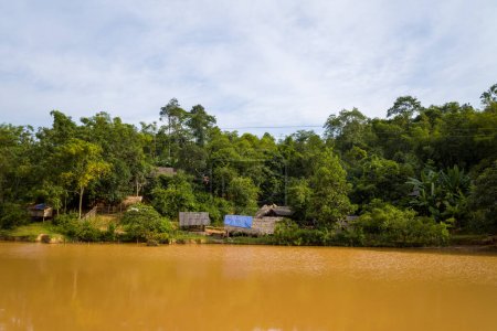 Cette photo de paysage a été prise, en Asie, au Vietnam, au Tonkin, vers Hanoi, à Mai Chau, en été. Nous voyons un étang boueux devant un village traditionnel, sous le soleil.