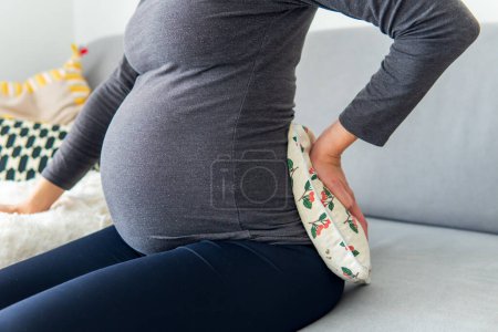 Foto de Mujer embarazada sentada cómodamente en el sofá y sosteniendo una almohada térmica terapéutica en su espalda baja mientras sostiene su vientre. Tratamiento del dolor de espalda durante el embarazo y uso de una almohadilla térmica para aliviar. - Imagen libre de derechos