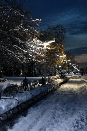 A winter and night city with people, benches, houses, buildings, white snow, bare trees, lanterns are located along the Dnipro River. Very unusual sections of the winter embankment of  Dnipro River in night city of Dnepropetrovsk/Dnipro, Ukraine. 