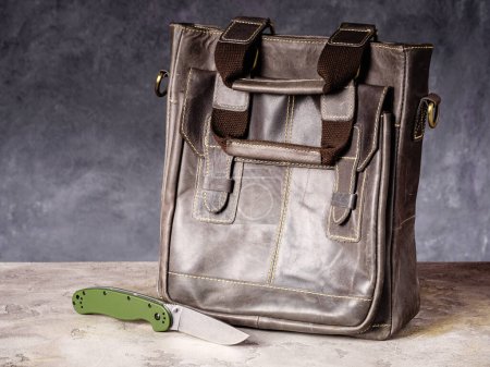 Foto de Green folding EDC knife near a leather business casual bag - Imagen libre de derechos
